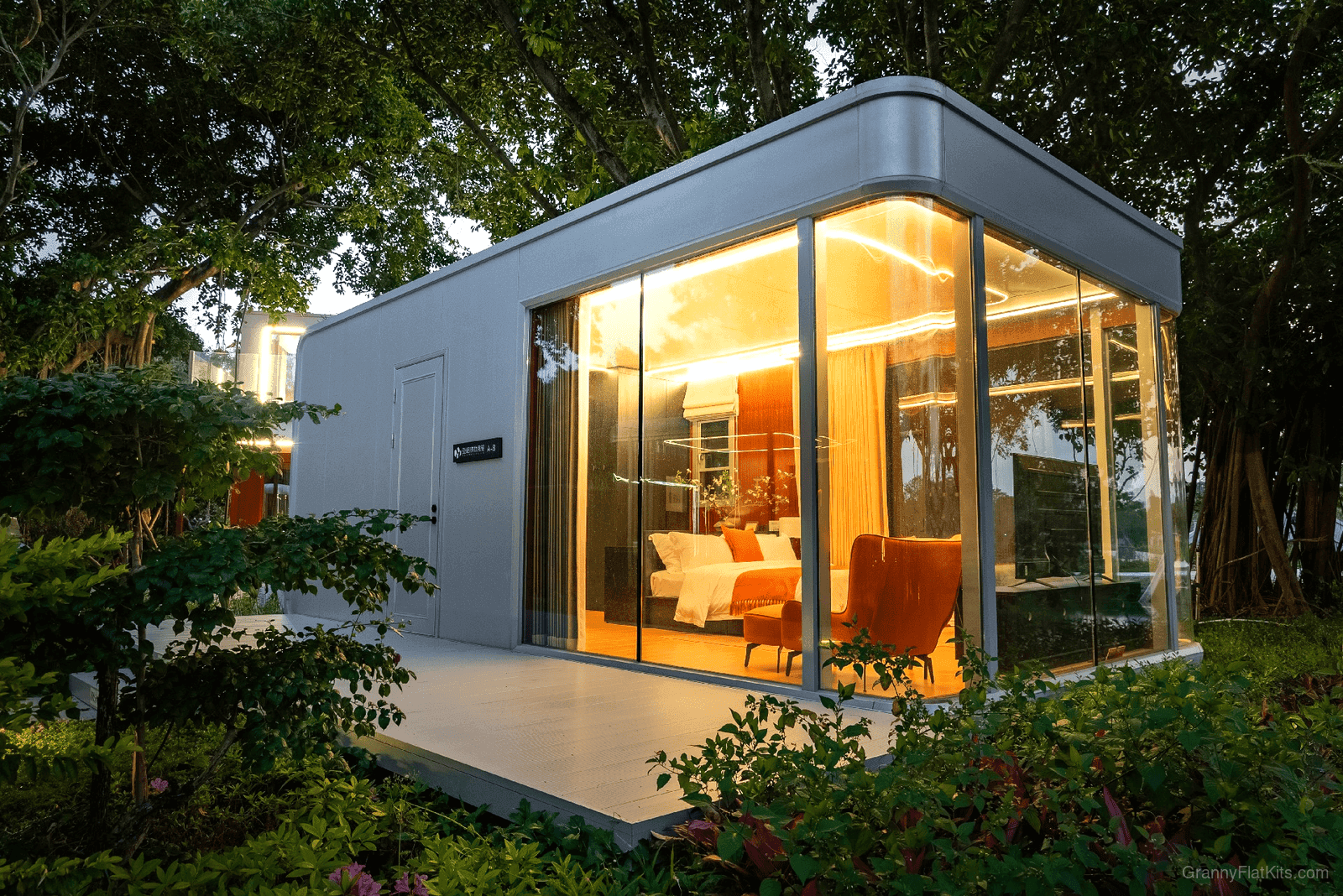 Compact prefab granny flat exterior at night with warm interior lighting through floor-to-ceiling glass walls